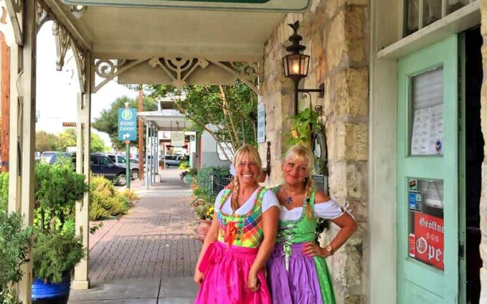 Two women in brightly colored German-style dresses stand before the fron door of Der Lindenbaum Restaurant in Texas.