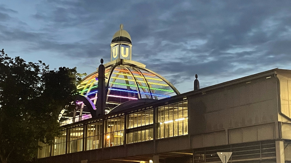 A picture of the Nollendorfplatz U-Bahn station at night, lit in the Pride colours