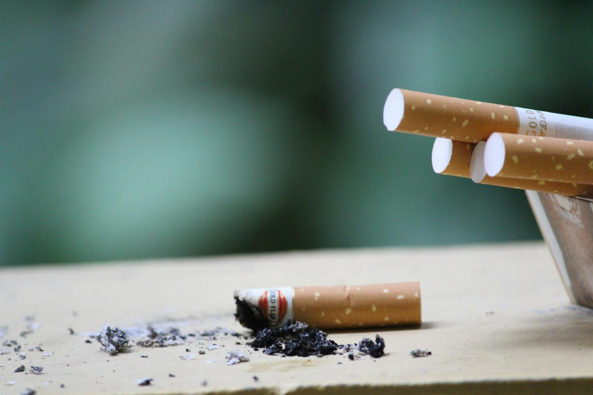 A burned cigarette on the table and more cigarettes in an ashtray next to it.