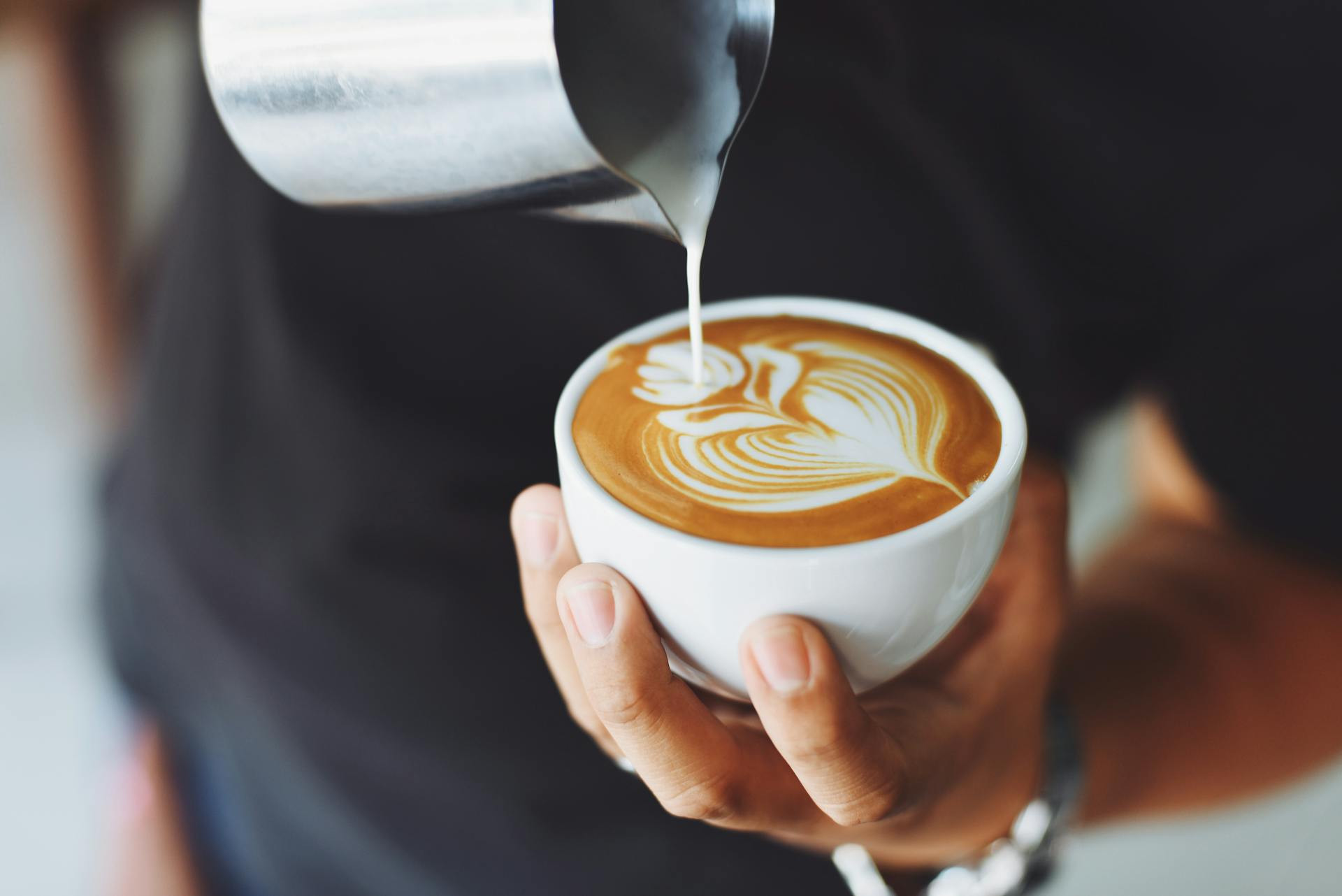 A close-up of a barrista drawing a flower with foamed milk in a cup of coffee.