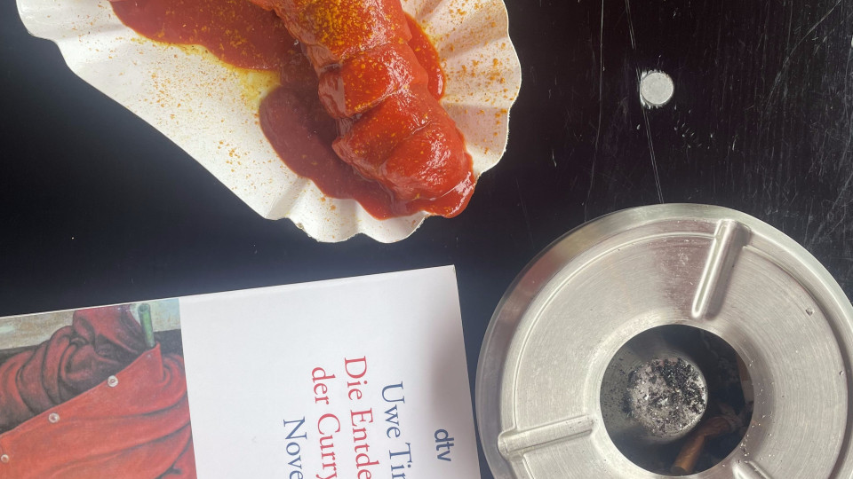 a picture of a plate of currywurst with Timm's book, 'The Invention of Currywurst' and an ashtray