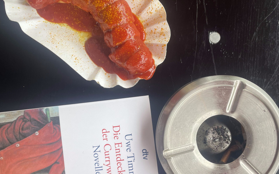 a picture of a plate of currywurst with Timm's book, 'The Invention of Currywurst' and an ashtray